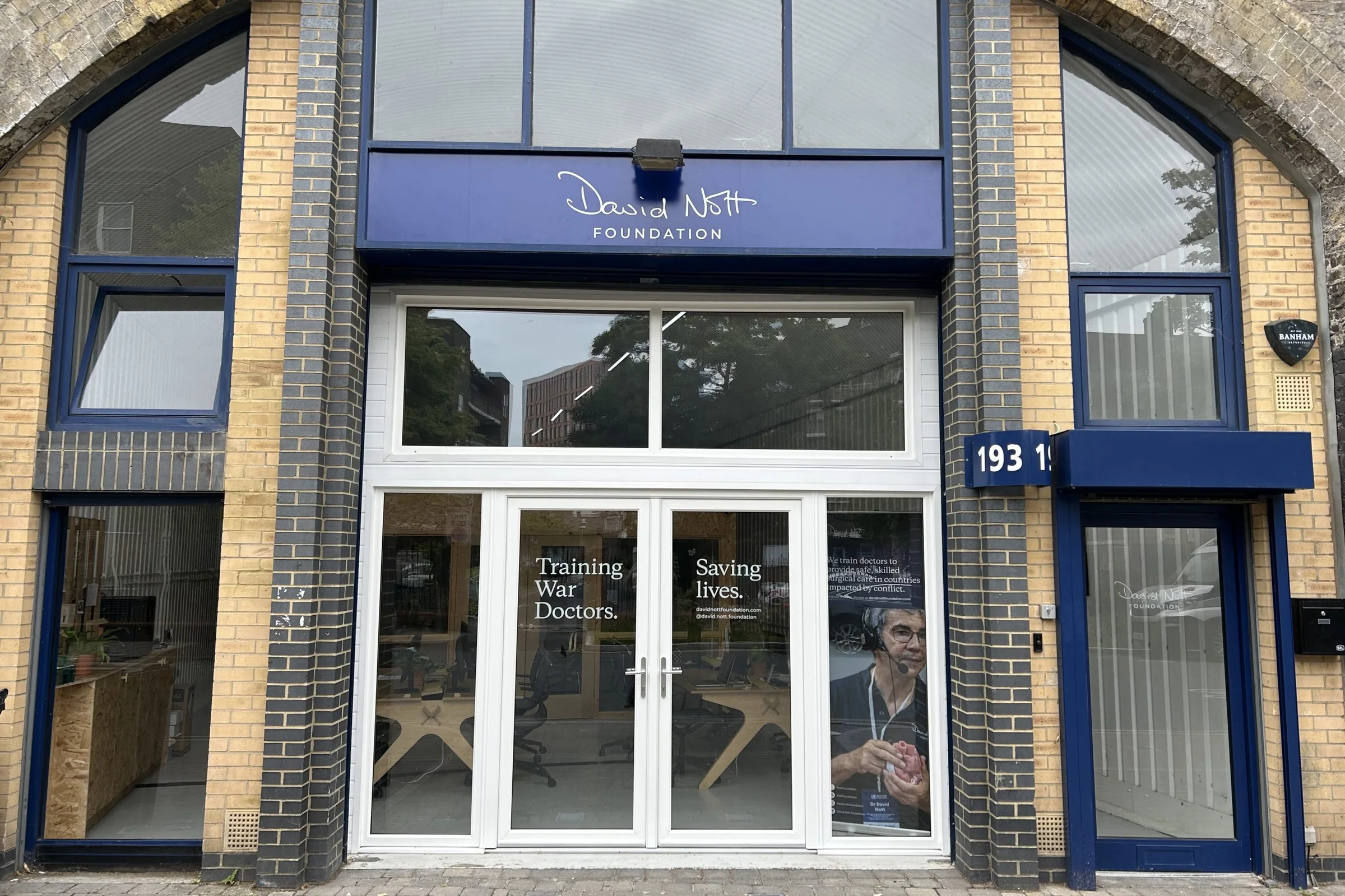 The David Nott Foundation office in a railway arch, with the signature logo on a blue fascia and ‘Training war doctors. Saving lives.’ vinyls on the glass doors